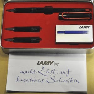K&ouml;nnte beinhalten: Ein schwarzes Lamy-Stiftset in einem rot ausgekleideten Metallkoffer. Das Set enth&auml;lt einen Stift, zwei Minen und eine kleine Schachtel Tintenpatronen. Das Lamy-Logo ist auf der Schachtel und dem Stift zu sehen. Ein Blatt Papier mit dem Lamy-Logo und Text befindet sich vor dem Koffer.