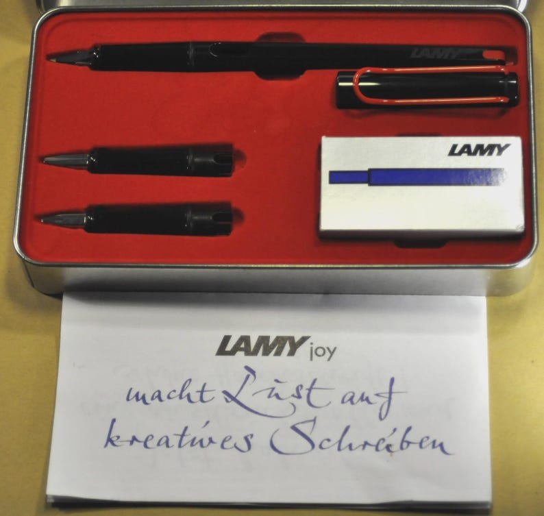 K&ouml;nnte beinhalten: Ein schwarzes Lamy-Stiftset in einem rot ausgekleideten Metallkoffer. Das Set enth&auml;lt einen Stift, zwei Minen und eine kleine Schachtel Tintenpatronen. Das Lamy-Logo ist auf der Schachtel und dem Stift zu sehen. Ein Blatt Papier mit dem Lamy-Logo und Text befindet sich vor dem Koffer.