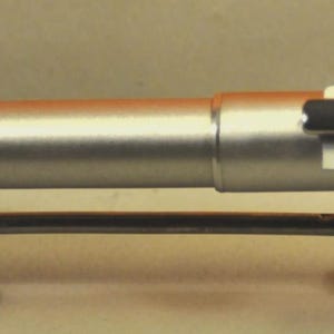 May include: A silver-colored pen with a black clip, resting on a decorative metal stand. The pen has a cylindrical body and appears to be made of metal. The stand has a horizontal bar supported by four spherical feet.