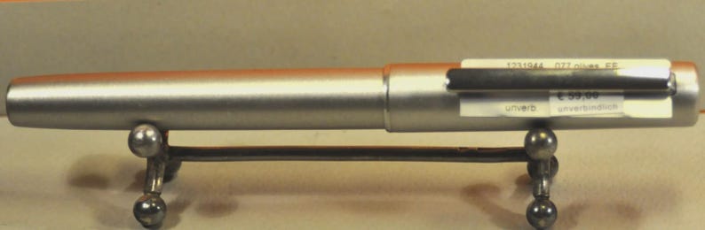 May include: A silver-colored pen with a black clip, resting on a decorative metal stand. The pen has a cylindrical body and appears to be made of metal. The stand has a horizontal bar supported by four spherical feet.