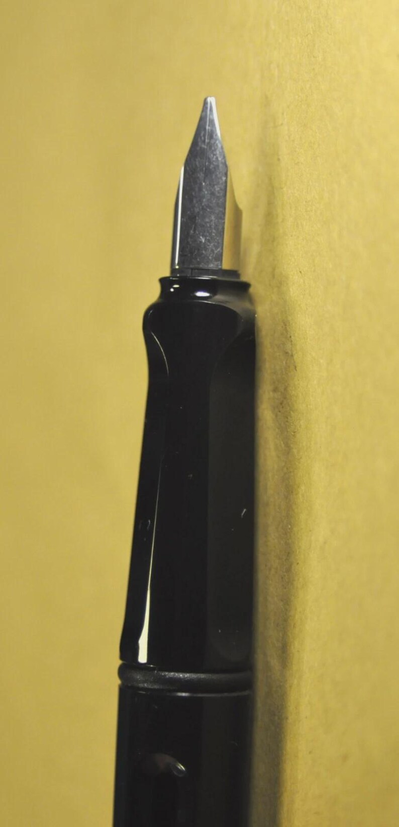 K&ouml;nnte beinhalten: Nahaufnahme eines schwarzen F&uuml;llfederhalters mit silberner Feder. Der Stift hat ein schlankes, modernes Design mit gl&auml;nzendem Finish. Die Feder ist spitz und scheint zum Schreiben bereit zu sein. Der Hintergrund ist einfarbig, ged&auml;mpftes Gelb.