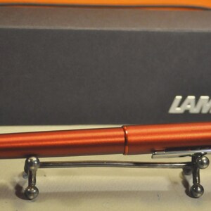 May include: A metallic copper-colored fountain pen with a silver-toned clip featuring the LAMY logo, resting on a small silver metal stand. Behind the pen sits a rectangular black box with the silver LAMY brand name printed on the front right side.