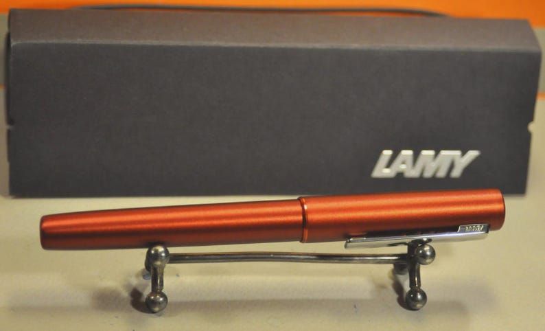 May include: A metallic copper-colored fountain pen with a silver-toned clip featuring the LAMY logo, resting on a small silver metal stand. Behind the pen sits a rectangular black box with the silver LAMY brand name printed on the front right side.
