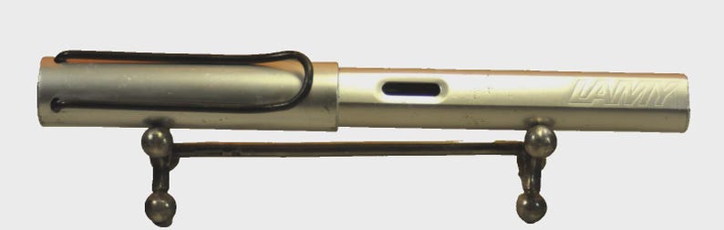 K&ouml;nnte beinhalten: Ein silberfarbener Lamy-F&uuml;llfederhalter mit matter Oberfl&auml;che, ausgestattet mit einem schwarzen Drahtclip an der Kappe und einem rechteckigen Sichtfenster f&uuml;r die Tinte am Schaft. Der Stift liegt waagerecht auf einem kleinen, dunklen Metallst&auml;nder.