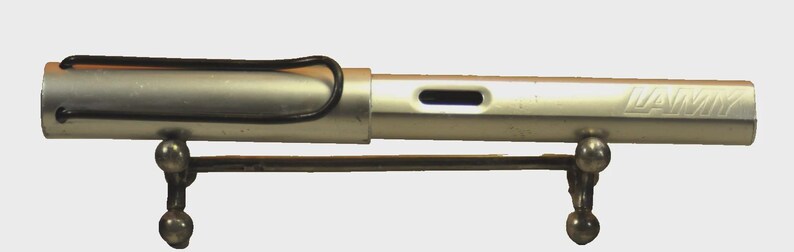 K&ouml;nnte beinhalten: Ein silberfarbener Lamy-F&uuml;llfederhalter mit matter Oberfl&auml;che, ausgestattet mit einem schwarzen Drahtclip an der Kappe und einem rechteckigen Sichtfenster f&uuml;r die Tinte am Schaft. Der Stift liegt waagerecht auf einem kleinen, dunklen Metallst&auml;nder.