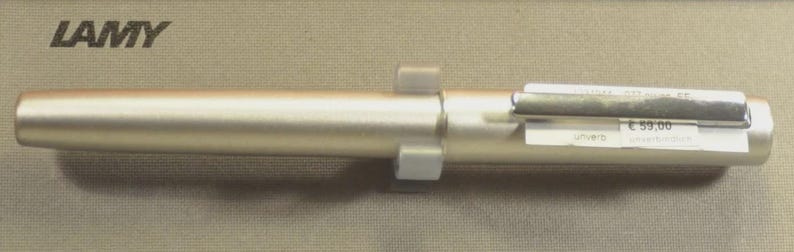 Może przedstawiać: Srebrny długopis LAMY z prostokątnym klipsem i białą metką z ceną. Długopis leży na jasnobrązowej powierzchni. Logo LAMY znajduje się w lewym g&oacute;rnym rogu.