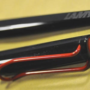 K&ouml;nnte beinhalten: Schwarzer Lamy-Stift mit rotem Clip und Akzenten. Der Stift hat einen schwarzen Schaft mit einem wei&szlig;en Streifen. Die Kappe hat einen roten Akzent und das Lamy-Logo ist sichtbar.