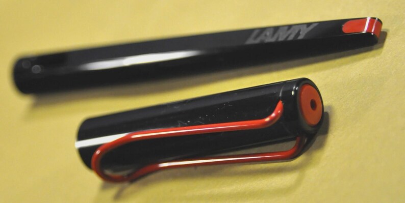 K&ouml;nnte beinhalten: Schwarzer Lamy-Stift mit rotem Clip und Akzenten. Der Stift hat einen schwarzen Schaft mit einem wei&szlig;en Streifen. Die Kappe hat einen roten Akzent und das Lamy-Logo ist sichtbar.