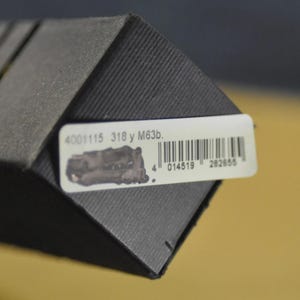 Puede incluir: Primer plano de una caja gris oscura texturizada con una etiqueta blanca. La etiqueta tiene un c&oacute;digo de barras, una peque&ntilde;a ilustraci&oacute;n y el texto "4001115 318 y M63b."