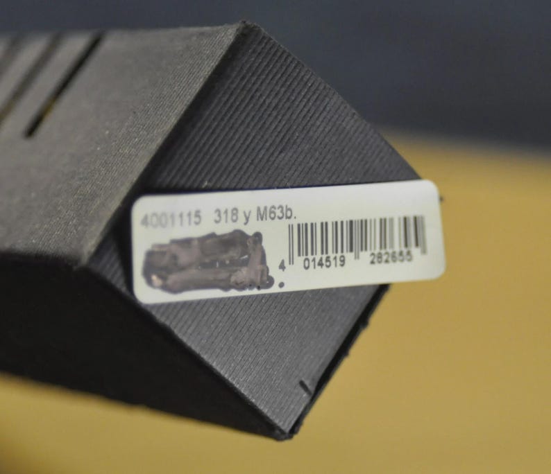 Puede incluir: Primer plano de una caja gris oscura texturizada con una etiqueta blanca. La etiqueta tiene un c&oacute;digo de barras, una peque&ntilde;a ilustraci&oacute;n y el texto "4001115 318 y M63b."