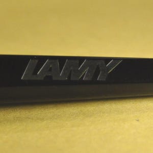 K&ouml;nnte beinhalten: Nahaufnahme eines schwarzen Stifts mit dem Wort "LAMY" auf der Seite. Der Stift hat einen leuchtend orangefarbenen Akzent an der Spitze. Der Stift befindet sich vor einem gelben Hintergrund.