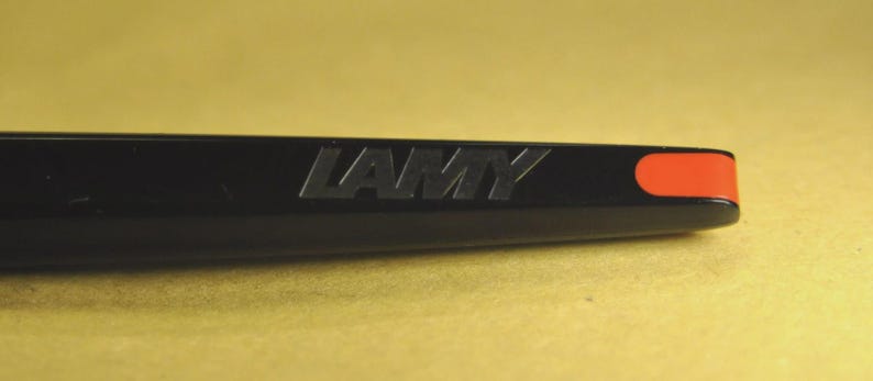 K&ouml;nnte beinhalten: Nahaufnahme eines schwarzen Stifts mit dem Wort "LAMY" auf der Seite. Der Stift hat einen leuchtend orangefarbenen Akzent an der Spitze. Der Stift befindet sich vor einem gelben Hintergrund.