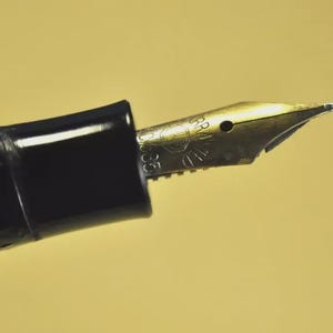 Puede incluir: Una pluma estilogr&aacute;fica negra con un plum&iacute;n de metal dorado. El plum&iacute;n muestra un logotipo circular grabado y el texto 'WARRANTED', '14K' y '585'. El cuerpo de la pluma est&aacute; hecho de un material negro liso y brillante con una secci&oacute;n de agarre estriada.