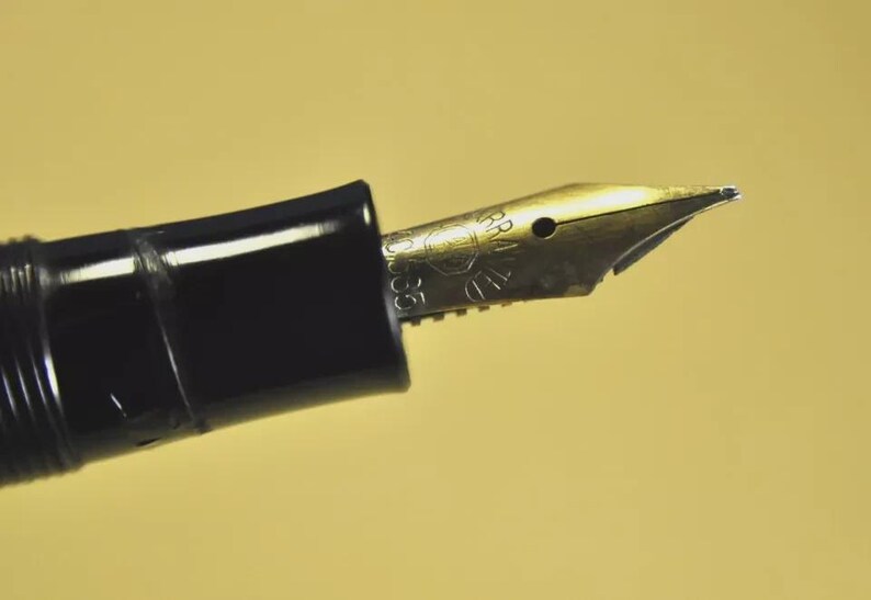 Puede incluir: Una pluma estilogr&aacute;fica negra con un plum&iacute;n de metal dorado. El plum&iacute;n muestra un logotipo circular grabado y el texto 'WARRANTED', '14K' y '585'. El cuerpo de la pluma est&aacute; hecho de un material negro liso y brillante con una secci&oacute;n de agarre estriada.