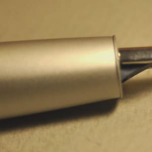 May include: Close-up of a silver fountain pen. The pen's body has a matte finish, and the nib is made of shiny metal. The nib is slightly curved and has a small slit. The pen is on a neutral-toned surface.