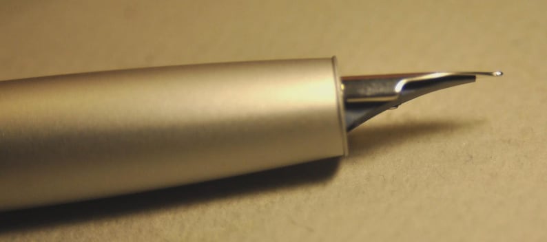 May include: Close-up of a silver fountain pen. The pen's body has a matte finish, and the nib is made of shiny metal. The nib is slightly curved and has a small slit. The pen is on a neutral-toned surface.
