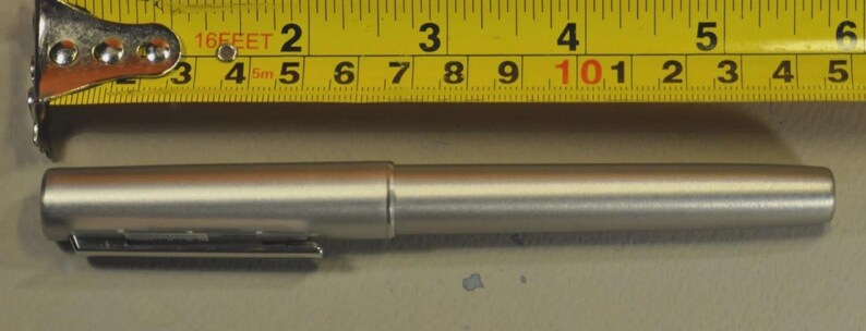 Może przedstawiać: Srebrny długopis z klipsem obok ż&oacute;łtej miarki. Długopis ma około 14 cm długości. Miarka pokazuje pomiary w stopach i calach.