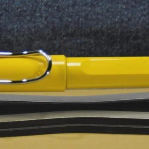 Puede incluir: Una pluma estilogr&aacute;fica de color amarillo brillante con clip y detalles plateados. La pluma se muestra horizontalmente sobre un soporte para bol&iacute;grafos blanco y negro. La pluma tiene la palabra "LAMY" grabada en el lateral.