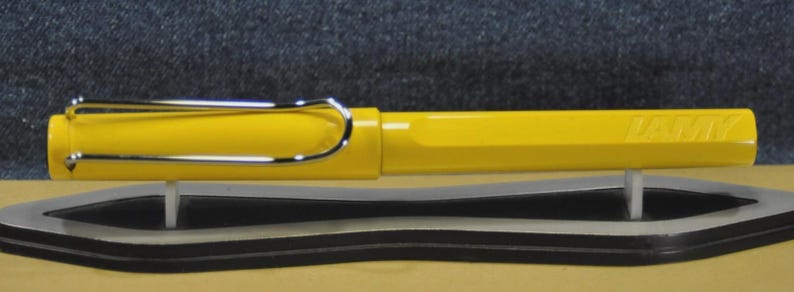 Puede incluir: Una pluma estilogr&aacute;fica de color amarillo brillante con clip y detalles plateados. La pluma se muestra horizontalmente sobre un soporte para bol&iacute;grafos blanco y negro. La pluma tiene la palabra "LAMY" grabada en el lateral.
