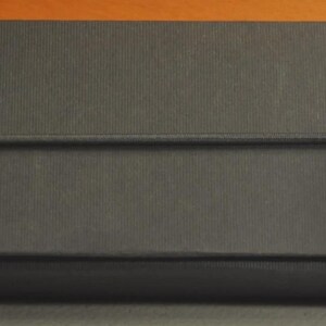 May include: A rectangular black cardboard box with a matte finish, featuring the white text LAMY printed on the lower right side. The box is secured by a thin black elastic band that wraps horizontally around the center of the lid.