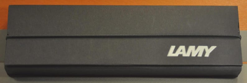May include: A rectangular black cardboard box with a matte finish, featuring the white text LAMY printed on the lower right side. The box is secured by a thin black elastic band that wraps horizontally around the center of the lid.