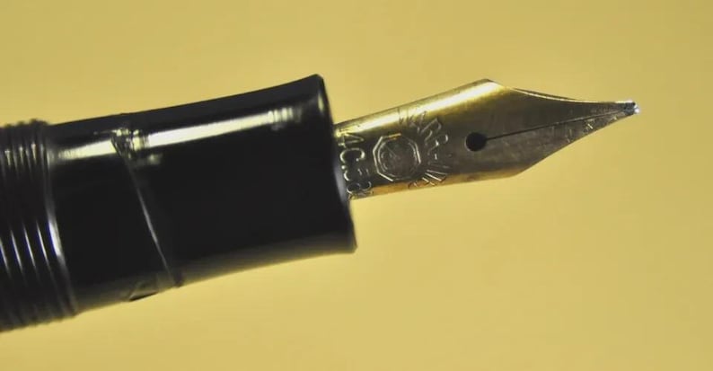 Puede incluir: Una pluma estilogr&aacute;fica negra con un plum&iacute;n de metal dorado. El plum&iacute;n presenta un logotipo circular grabado y el texto 4C58. La pluma tiene una secci&oacute;n de agarre negra lisa y un cuerpo acanalado, mostrando un dise&ntilde;o cl&aacute;sico de escritura.