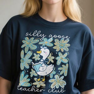 May include: Dark blue t-shirt with a playful illustration of a goose reading a book, set amongst blue and gold floral elements. The text reads "silly goose teacher club".