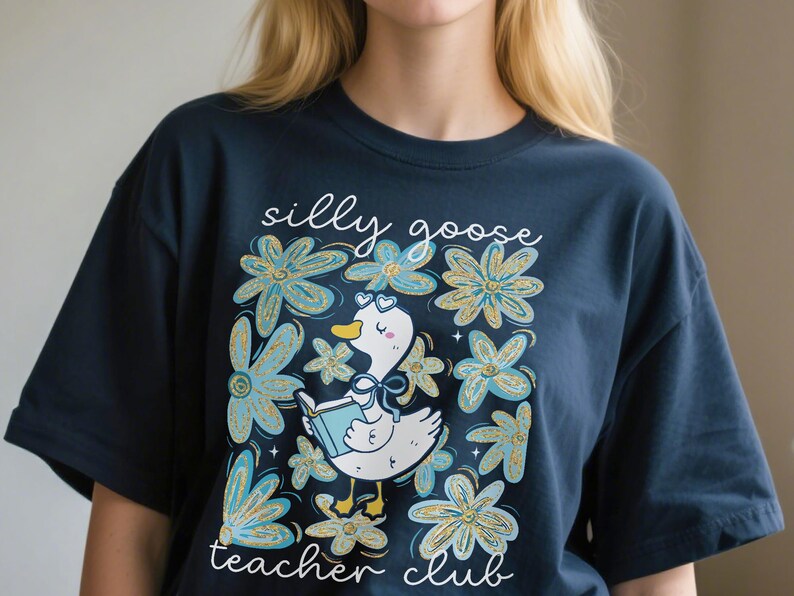May include: Dark blue t-shirt with a playful illustration of a goose reading a book, set amongst blue and gold floral elements. The text reads "silly goose teacher club".