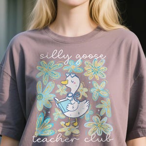 May include: Mauve t-shirt with a cartoon goose wearing heart-shaped glasses and reading a book, surrounded by blue and yellow flowers. The text "silly goose teacher club" is above and below the goose.
