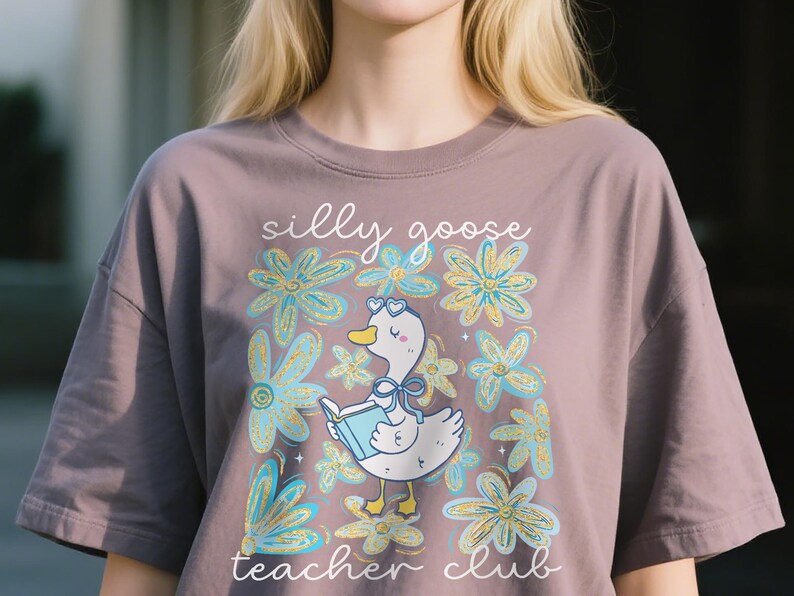 May include: Mauve t-shirt with a cartoon goose wearing heart-shaped glasses and reading a book, surrounded by blue and yellow flowers. The text "silly goose teacher club" is above and below the goose.