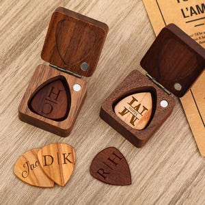Puede incluir: Cajas de púas de guitarra de madera con púas personalizadas. Las cajas son cuadradas con un recorte en forma de corazón en la tapa. Las púas están grabadas con iniciales y nombres. Las púas son de madera y de varios tonos marrones.