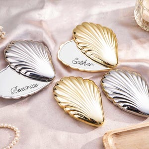 May include: Four seashell-shaped place card holders, two gold and two silver, with scalloped edges. Each holder has a white, heart-shaped base. Two bases have names, "Beatrice" and "Esther", written in cursive. A lit candle and decorative flowers are in the background.