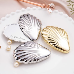 May include: A collection of shell-shaped compact mirrors in silver and gold tones, arranged on a white, scalloped plate. One mirror is engraved with the name "Andrea". Faux pearls are scattered around the mirrors, and makeup brushes are visible in the background.