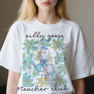 May include: White t-shirt with a playful design. It features a cartoon goose reading a book, surrounded by blue and gold flowers. The text "silly goose teacher club" is written above and below the goose illustration.