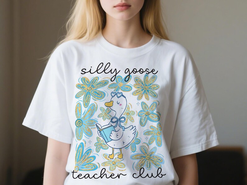 May include: White t-shirt with a playful design. It features a cartoon goose reading a book, surrounded by blue and gold flowers. The text "silly goose teacher club" is written above and below the goose illustration.