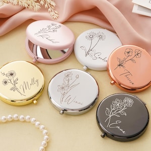 以下が含まれることがあります： ピンク、ゴールド、白、シルバー、銅、黒など、さまざまな色のパーソナライズされたコンパクトミラーのコレクション。各ミラーには花の模様と名前が特徴です。丸いミラーは開き、反射面が現れます。