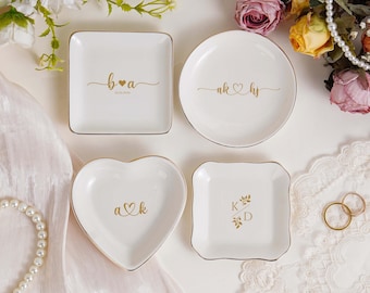 Pode incluir: Quatro pratos de cerâmica brancos para anéis com borda dourada. Um prato quadrado tem as iniciais "B & a" com uma data. Um prato redondo tem "ak ♡ hs". Um prato em forma de coração tem "a ♡ k". Um prato quadrado tem "K & D".