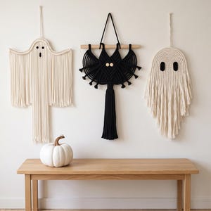 May include: Three macrame wall hangings featuring two off-white ghost shapes with dark eyes and mouths, and one central black bat shape with wooden bead eyes, all suspended by cords or a wooden dowel against a plain wall.