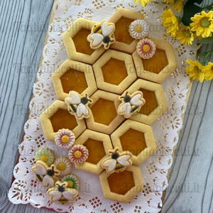 Può includere: Una collezione di biscotti esagonali con centri di marmellata arancione disposti a nido d'ape. I biscotti sono decorati con piccole glasse a forma di ape bianca e gialla e accenti floreali colorati su un centrino di pizzo bianco.