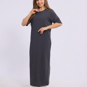 May include: A dark grey, floor-length dress with short sleeves. The dress has a simple, round neckline and a straight silhouette. The model is wearing white trainers. The dress is made of a soft, comfortable fabric.