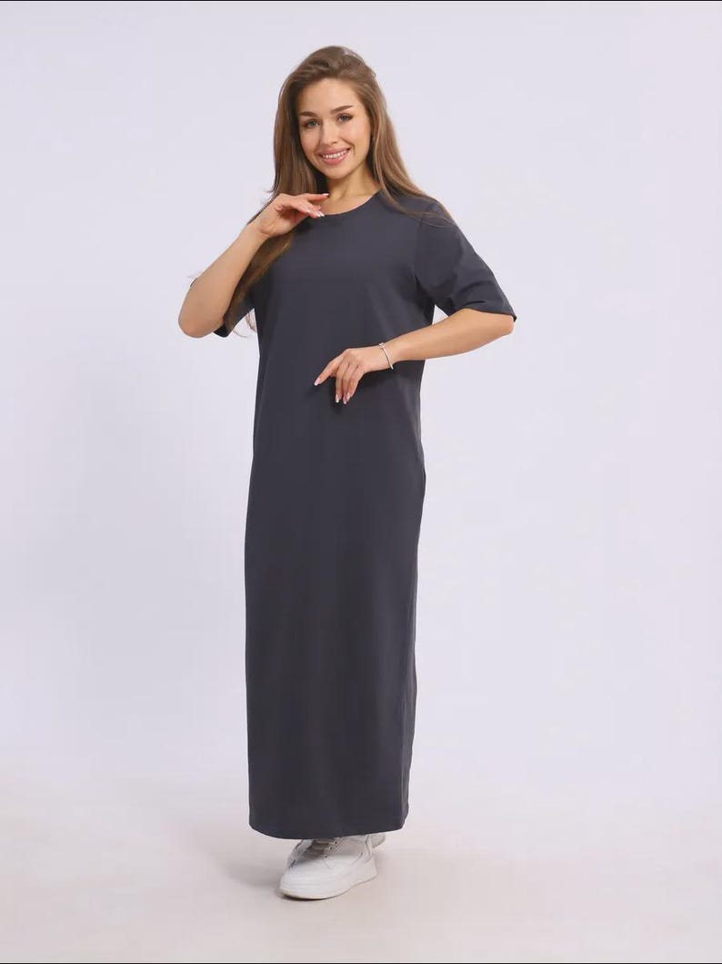 May include: A dark grey, floor-length dress with short sleeves. The dress has a simple, round neckline and a straight silhouette. The model is wearing white trainers. The dress is made of a soft, comfortable fabric.