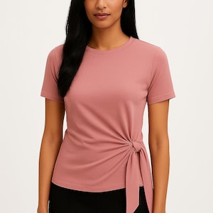 May include: A dusty rose-colored short-sleeved t-shirt with a round neckline. The shirt features a knotted detail on the left side. The model is wearing black pants. This top is suitable for casual wear.