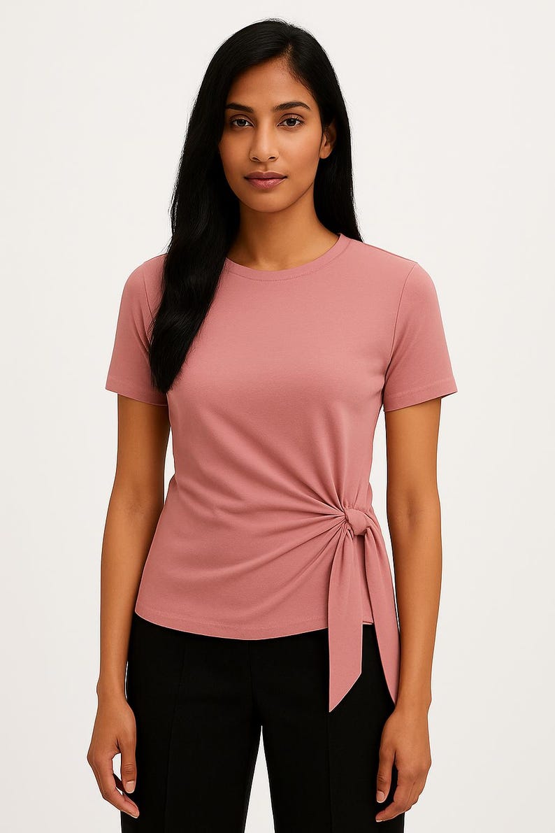 May include: A dusty rose-colored short-sleeved t-shirt with a round neckline. The shirt features a knotted detail on the left side. The model is wearing black pants. This top is suitable for casual wear.