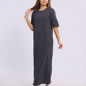 May include: A dark grey maxi dress with a round neck and short sleeves. The dress is worn with white trainers. The image is taken in a studio with a white backdrop.