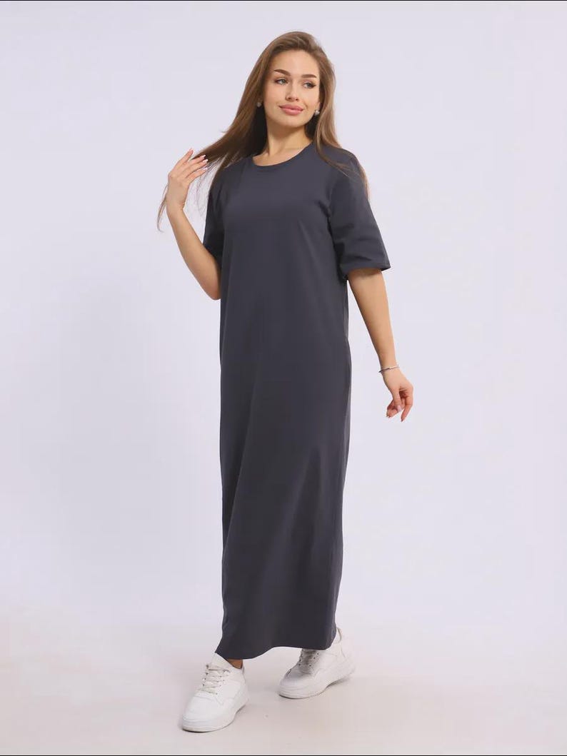 May include: A dark grey maxi dress with a round neck and short sleeves. The dress is worn with white trainers. The image is taken in a studio with a white backdrop.