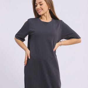 May include: A charcoal grey, short-sleeved dress with a round neckline. The dress has a simple, minimalist design and falls to the mid-calf. The model is wearing a silver bracelet and earrings. The dress is made of a soft, flowing fabric.