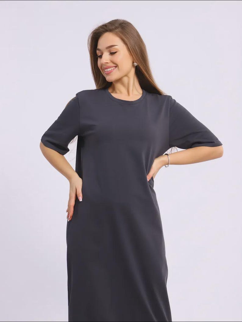 May include: A charcoal grey, short-sleeved dress with a round neckline. The dress has a simple, minimalist design and falls to the mid-calf. The model is wearing a silver bracelet and earrings. The dress is made of a soft, flowing fabric.