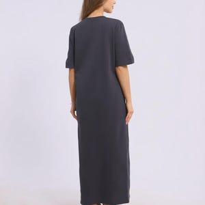May include: A dark grey maxi dress with short sleeves. The dress has a simple, relaxed fit and reaches the ankles. The model is wearing white trainers. The dress is made of a soft, comfortable material.