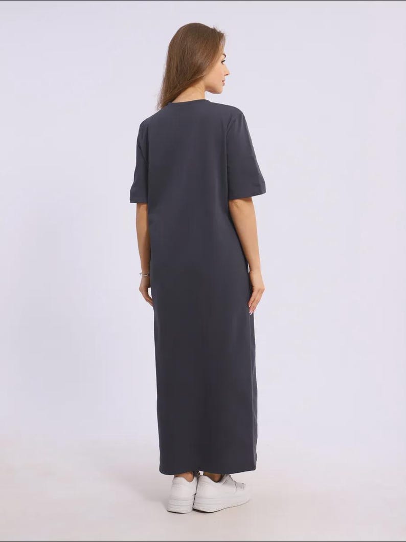 May include: A dark grey maxi dress with short sleeves. The dress has a simple, relaxed fit and reaches the ankles. The model is wearing white trainers. The dress is made of a soft, comfortable material.