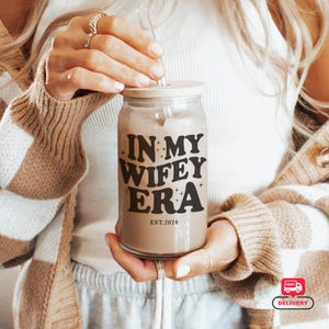 Puede incluir: Un vaso de vidrio transparente con tapa de madera y pajita de vidrio. El vaso está lleno de un líquido marrón claro y presenta el texto "IN MY WIFEY ERA" y "EST.2024" en negro. El vaso es sostenido por una persona.